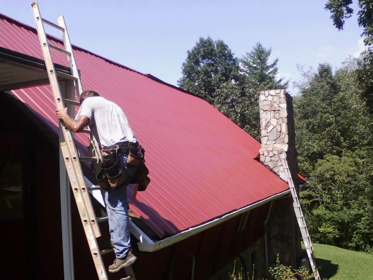 Roofer on a ladder beside a red metal roof and stone chimney — Soffit & Fascia Repair by Roofing Brothers in Callaway
